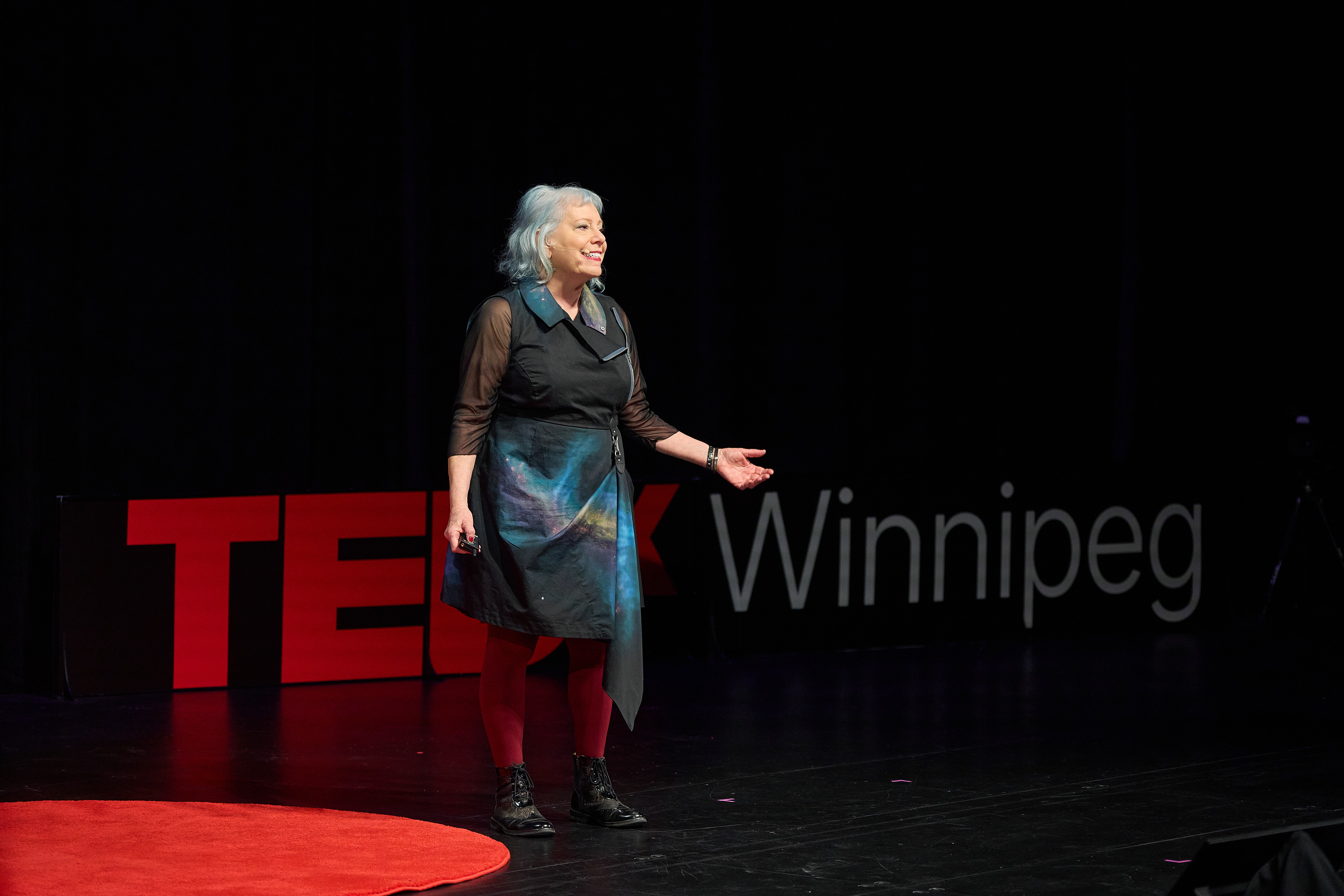 A speaker presents from the red circle carpet on the TEDxWinnipeg stage, with the event’s oversized letters behind her
